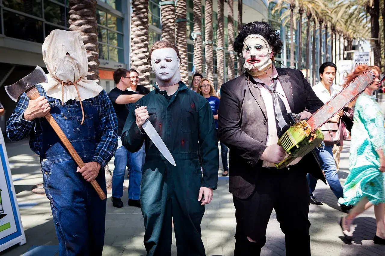 Trzech mężczyzn w strojach znanych postaci z horrorów: jeden z workiem na głowie i siekierą, drugi jako Michael Myers z nożem, trzeci jako Leatherface z piłą łańcuchową. Groźny slasher na konwencie.