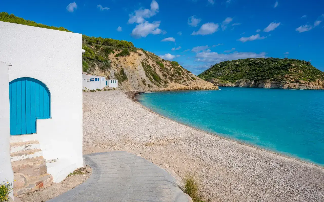 Casa blanca con puerta azul en la playa de guijarros de Vall d'Ebo, con aguas turquesas y colinas verdes.