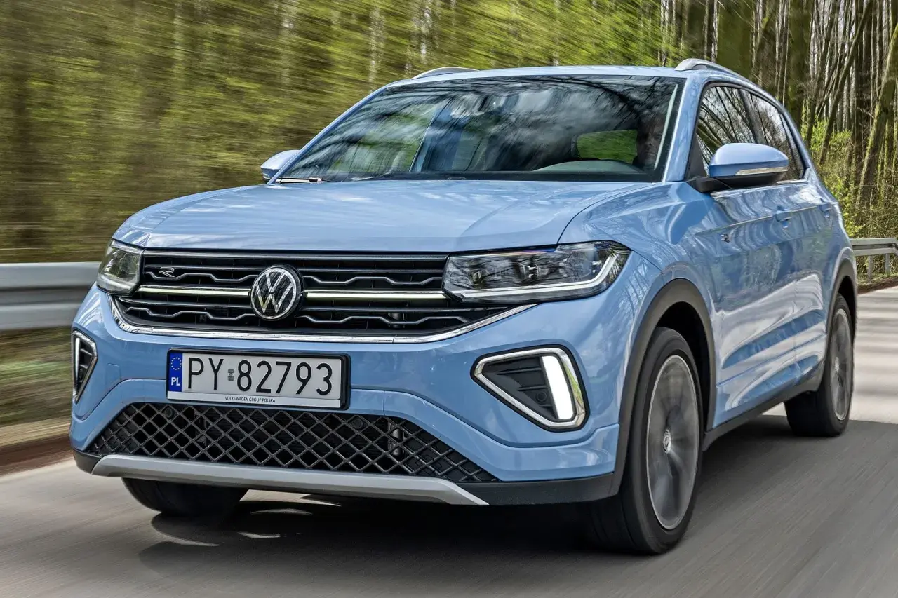 Niebieski VW T-Cross pędzi leśną drogą, odbijając drzewa w szybie.
