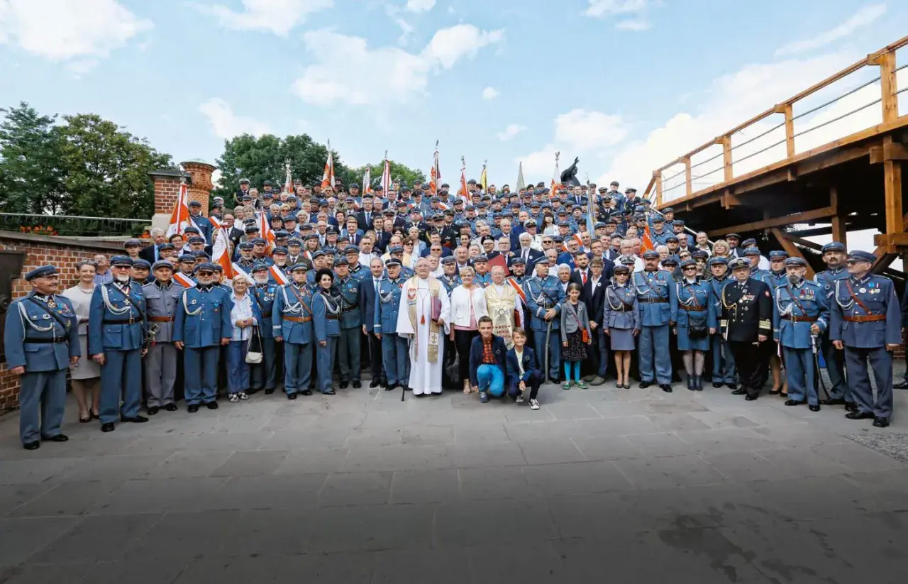 Grupa osób w historycznych mundurach, przypominających przedwojenne stopnie wojskowe, pozuje do zdjęcia na tle ceglanej budowli i drewnianej konstrukcji.