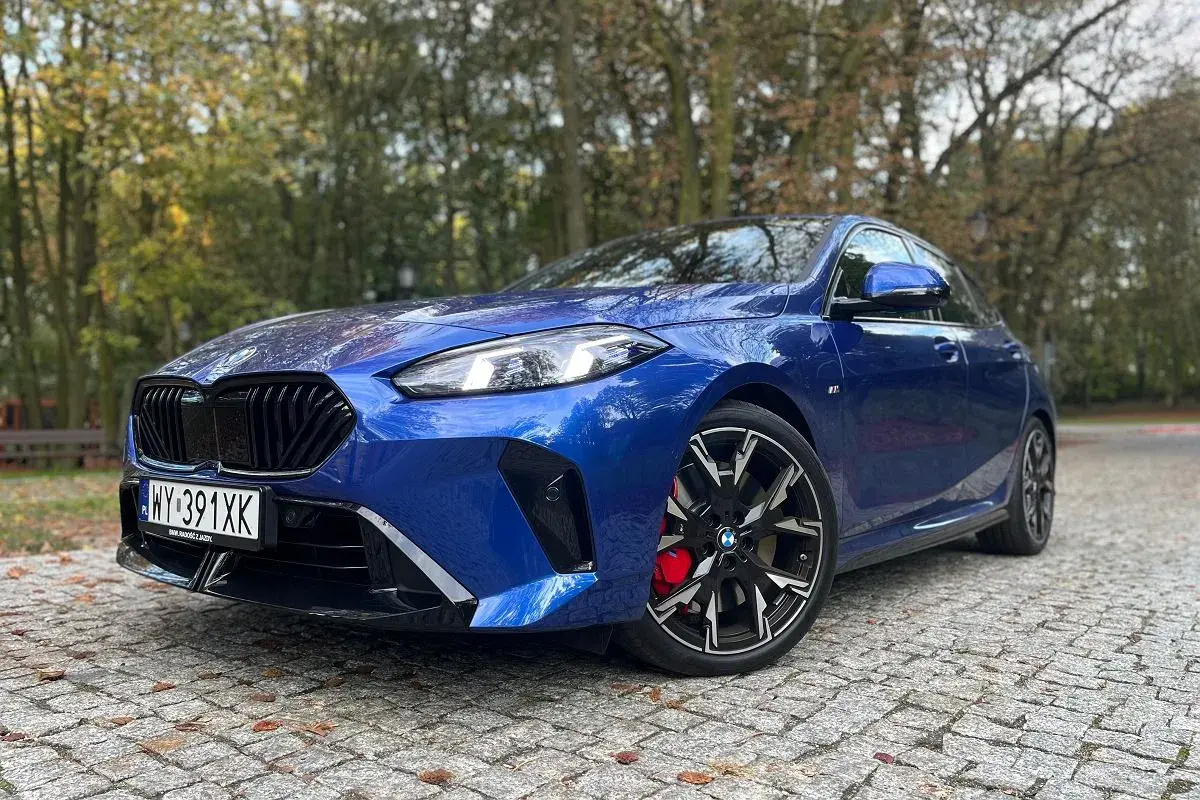 Błękitne BMW 235i z czarnymi felgami i czerwonymi zaciskami hamulcowymi zaparkowane na bruku wśród jesiennych drzew.