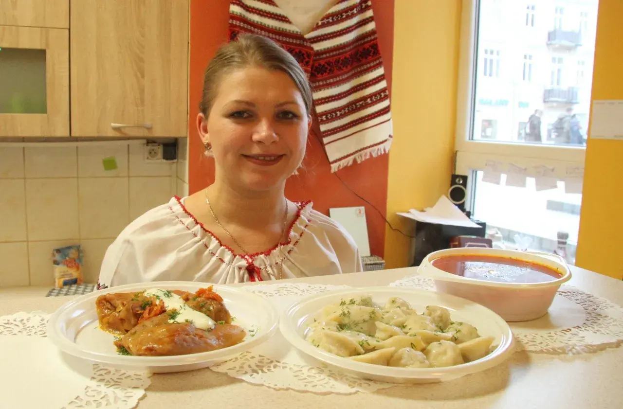 Uśmiechnięta kobieta w tradycyjnej koszuli prezentuje dania: gołąbki, pierogi i zupę. Pyszny, domowy obiad jak u Tarasa Bulby.