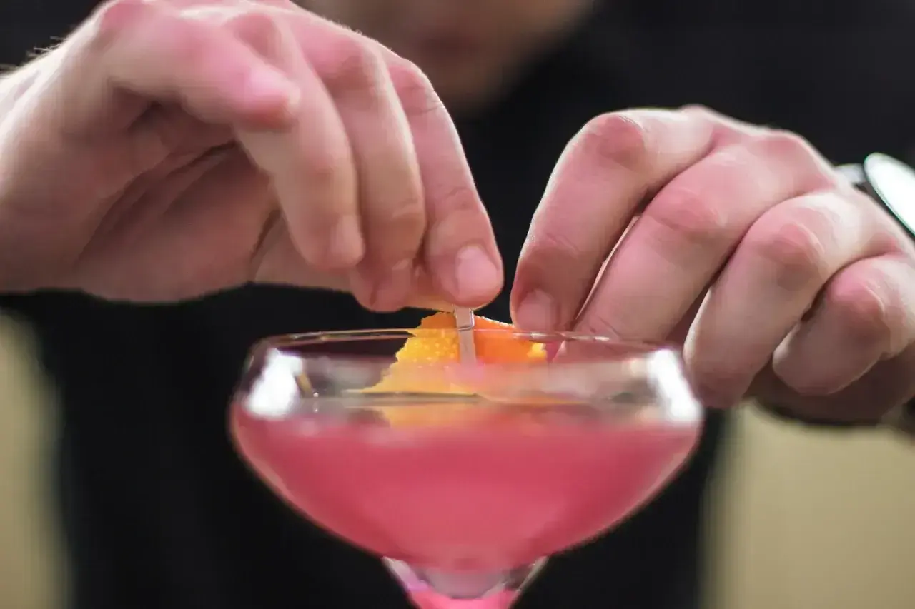 Dłonie barmana ozdabiają drinka skórką pomarańczy. To klasyczny cosmopolitan, gotowy na wieczór w wielkim mieście.