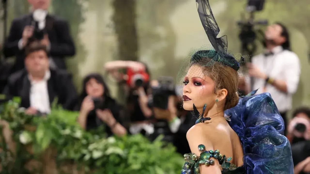 Una modelo con un elaborado atuendo de **Met Gala theme**, con velo y maquillaje dramático, posa rodeada de fotógrafos y vegetación.