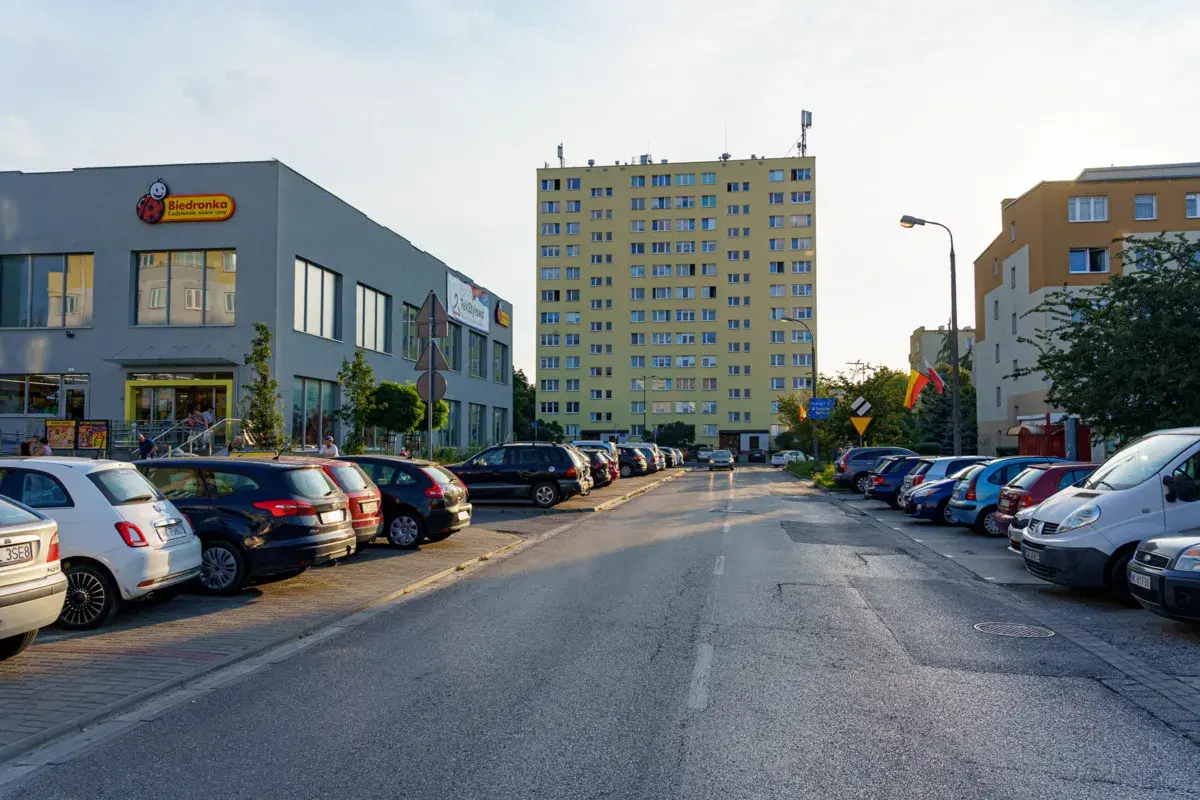 Uliczka z zaparkowanymi samochodami obok sklepu Biedronka, gdzie logo przypomina uśmiechniętego ursusa niedźwiadka. W tle wysoki, żółty blok mieszkalny.