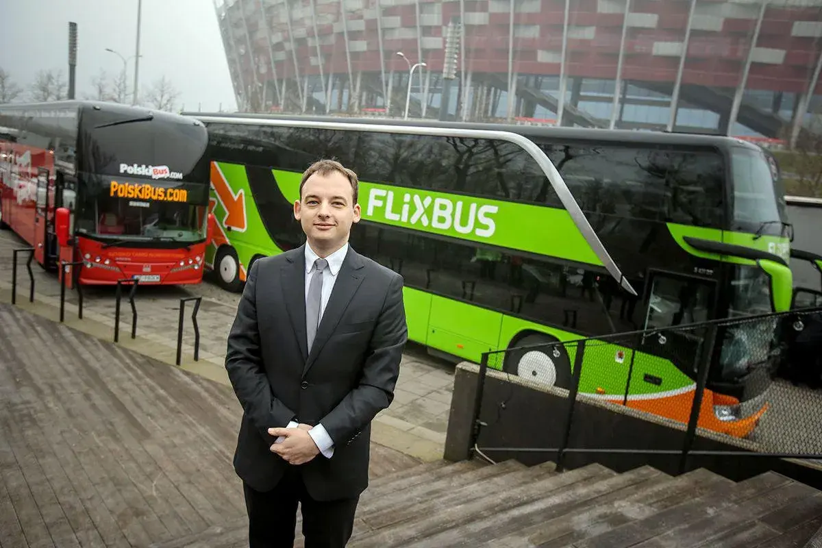 Mężczyzna w garniturze stoi przed zielonym autokarem Flixbus i czerwonym autokarem Polskibus.