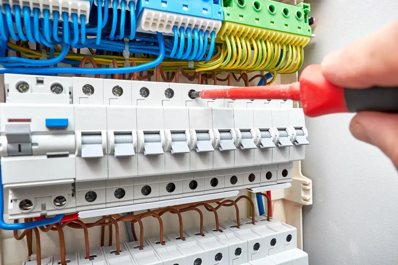 Ręka z czerwonym śrubokrętem przykręca przewody w skrzynce elektrycznej. Trwa wymiana instalacji elektrycznej.