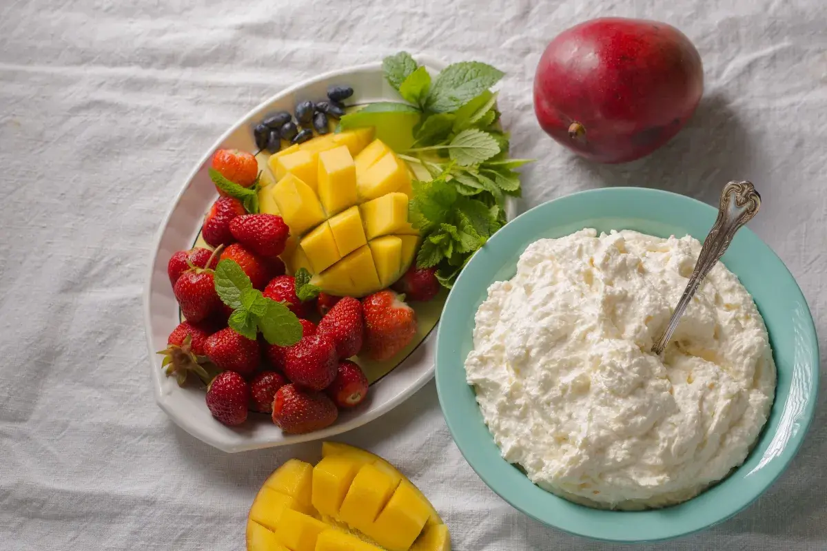 Un piatto di fragole, mango e mirtilli accanto a una ciotola di cosa sono i fiocchi di latte, con un mango intero sullo sfondo.