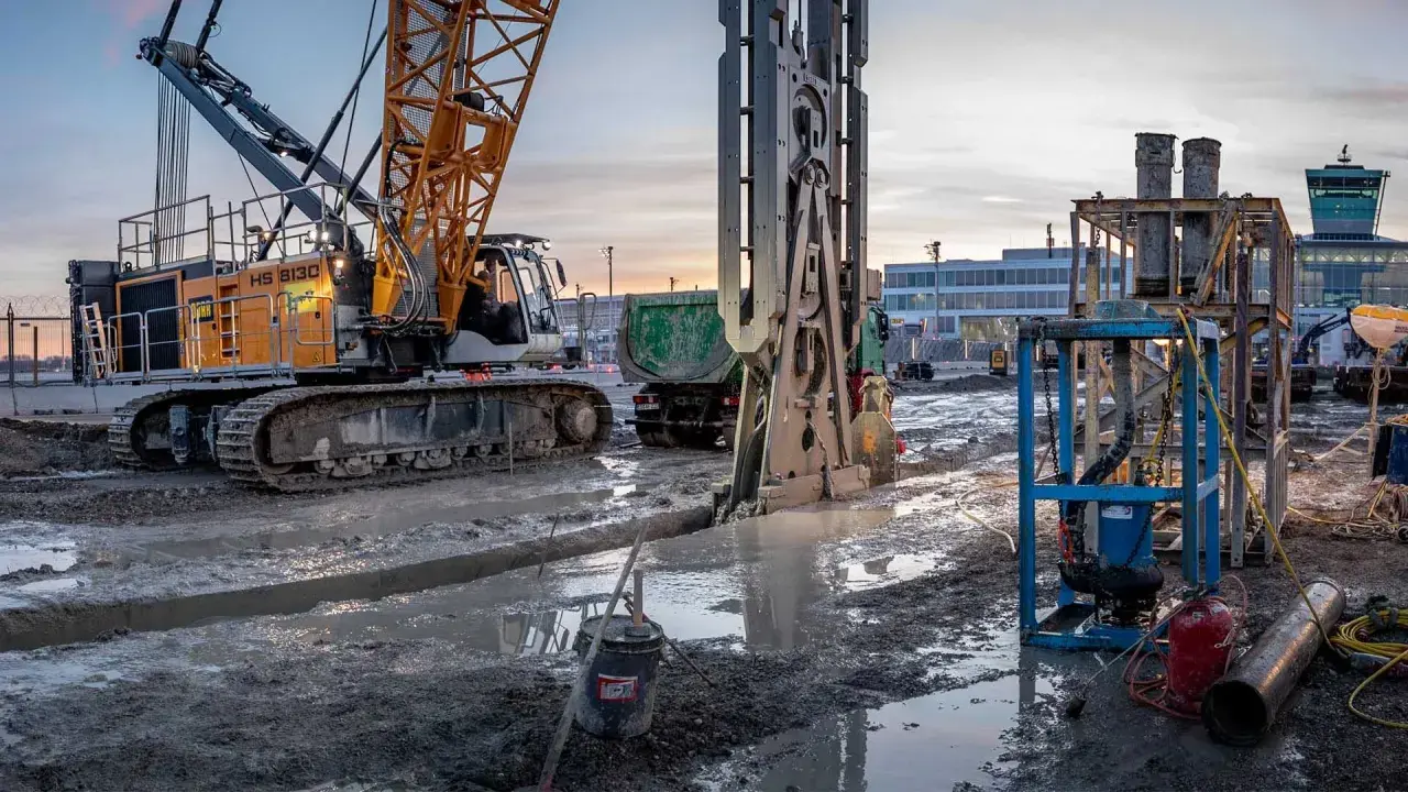 Baustelle bei Dämmerung: Kran und Bohranlage im Einsatz für die Schlitzwand Herstellung. Schlammige Gräben und schwere Maschinen prägen das Bild.
