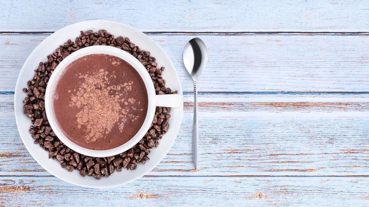 Tazza di cioccolata calda con cacao amaro in polvere e chicchi di cioccolato, su un piatto bianco, accanto a un cucchiaino. Le proprietà del cacao amaro sono note per il benessere.
