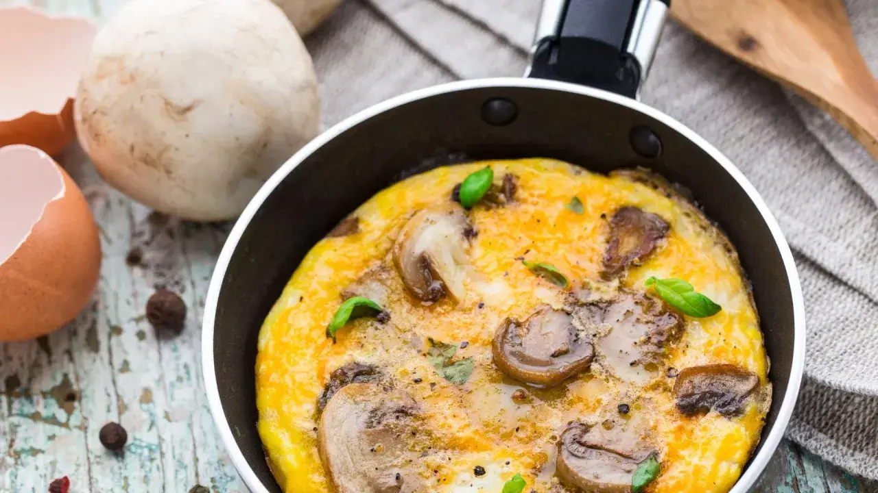Pyszny omlet z pieczarkami, świeżo usmażony na patelni, z dodatkiem bazylii. Obok skorupki jajka i grzyby.