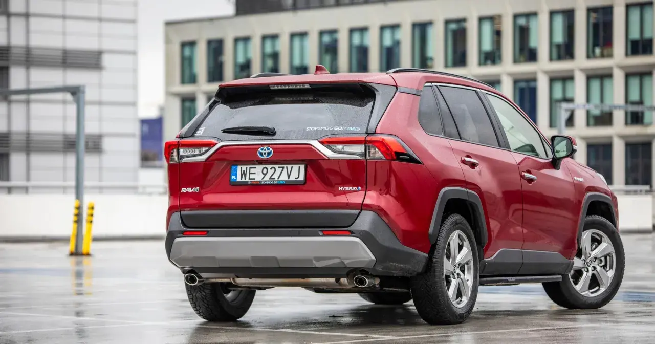 Czerwony Toyota RAV4 2020 Hybrid na mokrym parkingu, z nowoczesnym budynkiem w tle.