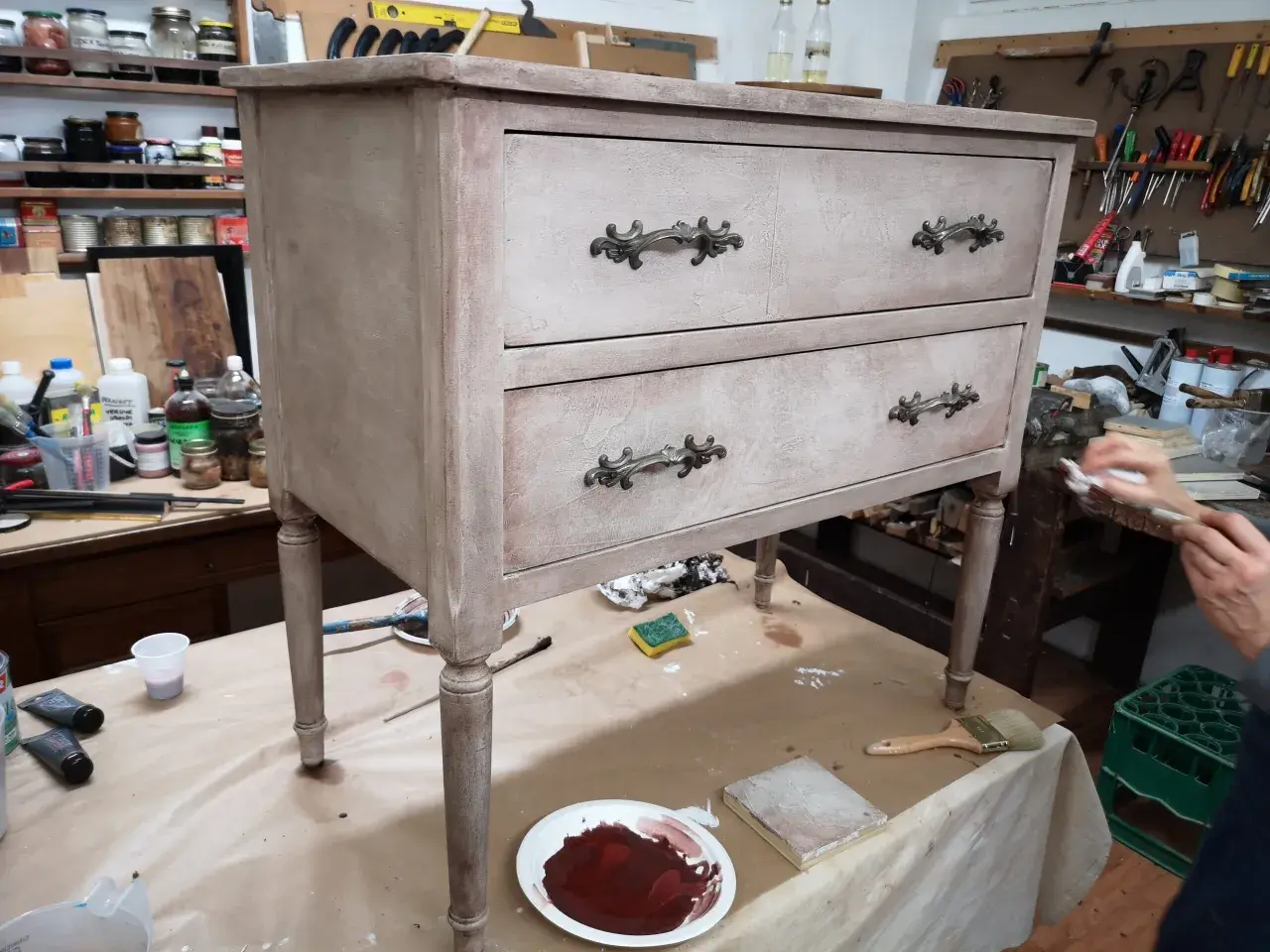 Un mobile antico in fase di restauro, con cassetti e pomelli decorati. Attrezzi e materiali da lavoro circondano il pezzo.