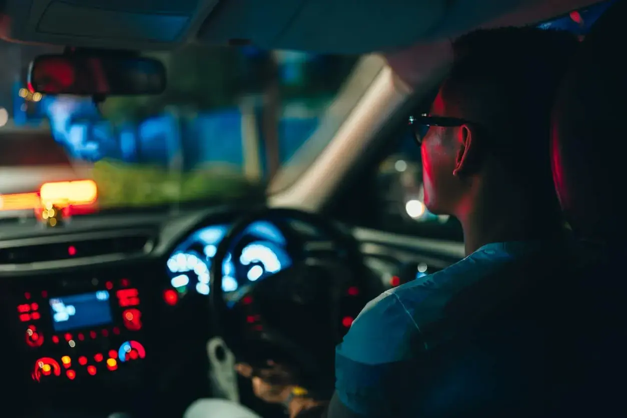 Un hombre con gafas conduce de noche, con el tablero iluminado en azul y rojo.
