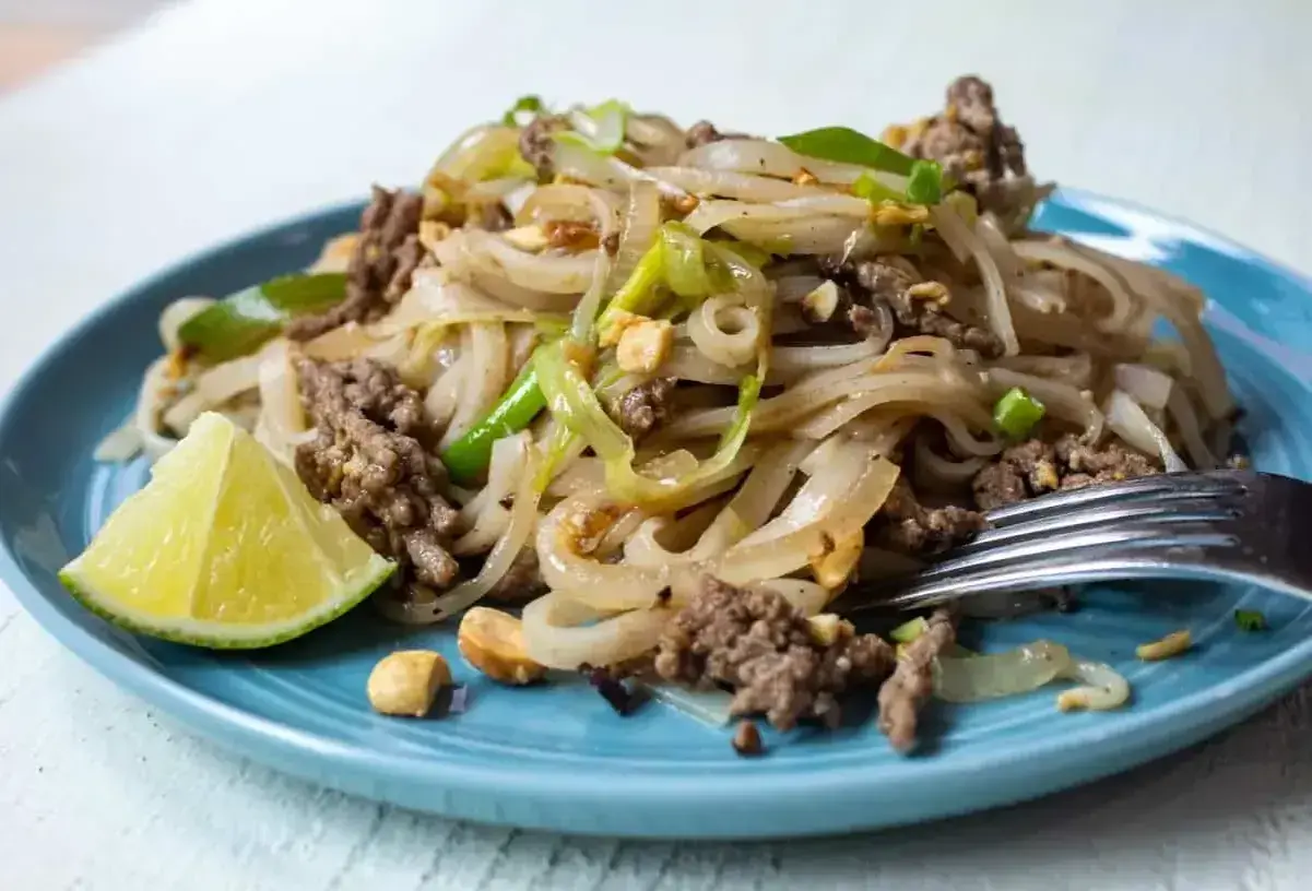 Pyszny pad thai przepis na talerzu: makaron ryżowy z mielonym mięsem, cebulą, orzeszkami ziemnymi i limonką.