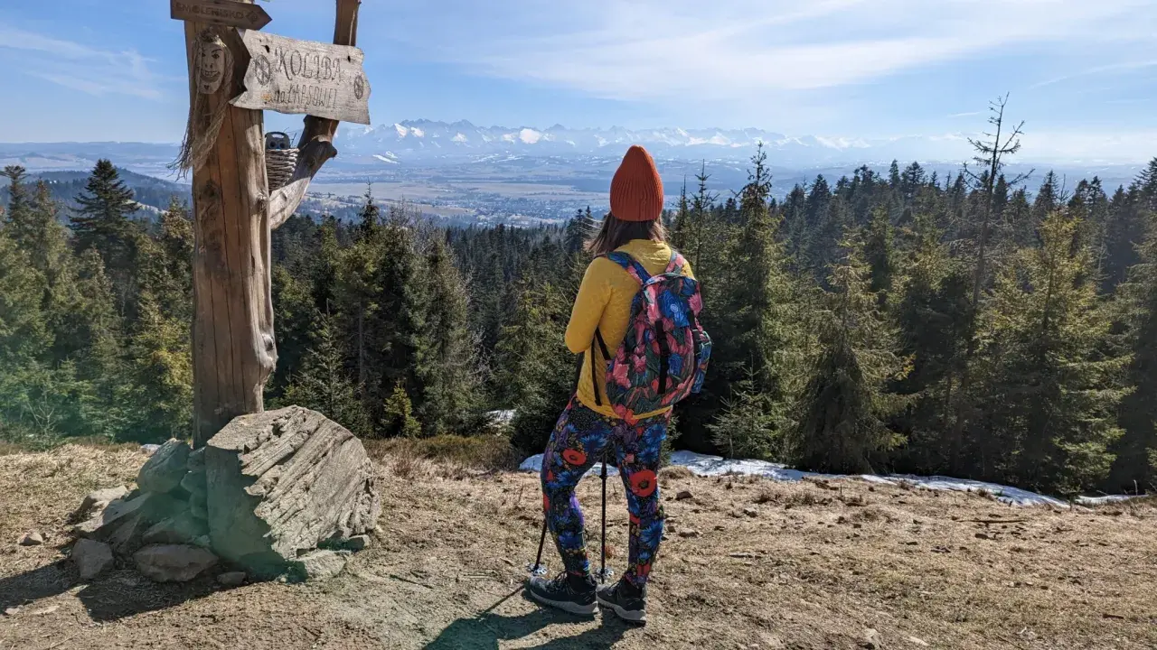 Widok na Tatry z drewnianego słupa z napisem "Koliba na Łapsowej Polanie". Turystka w kolorowych legginsach podziwia panoramę.