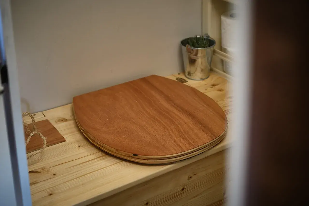 Un siège en bois pour toilettes sèches de camping-car, avec un petit seau de plantes et du papier toilette à proximité.