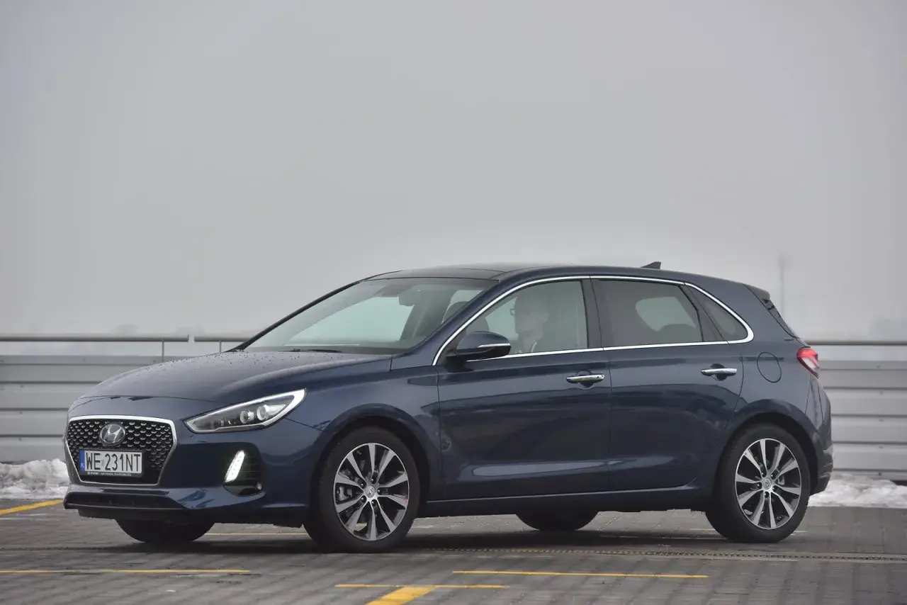 Ciemnoniebieski Hyundai i30 na parkingu. Kierowca w okularach siedzi za kierownicą, gotowy do wymiany żarówki.