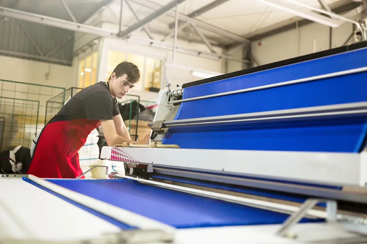 Un homme en tablier rouge travaille sur une machine textile bleue.