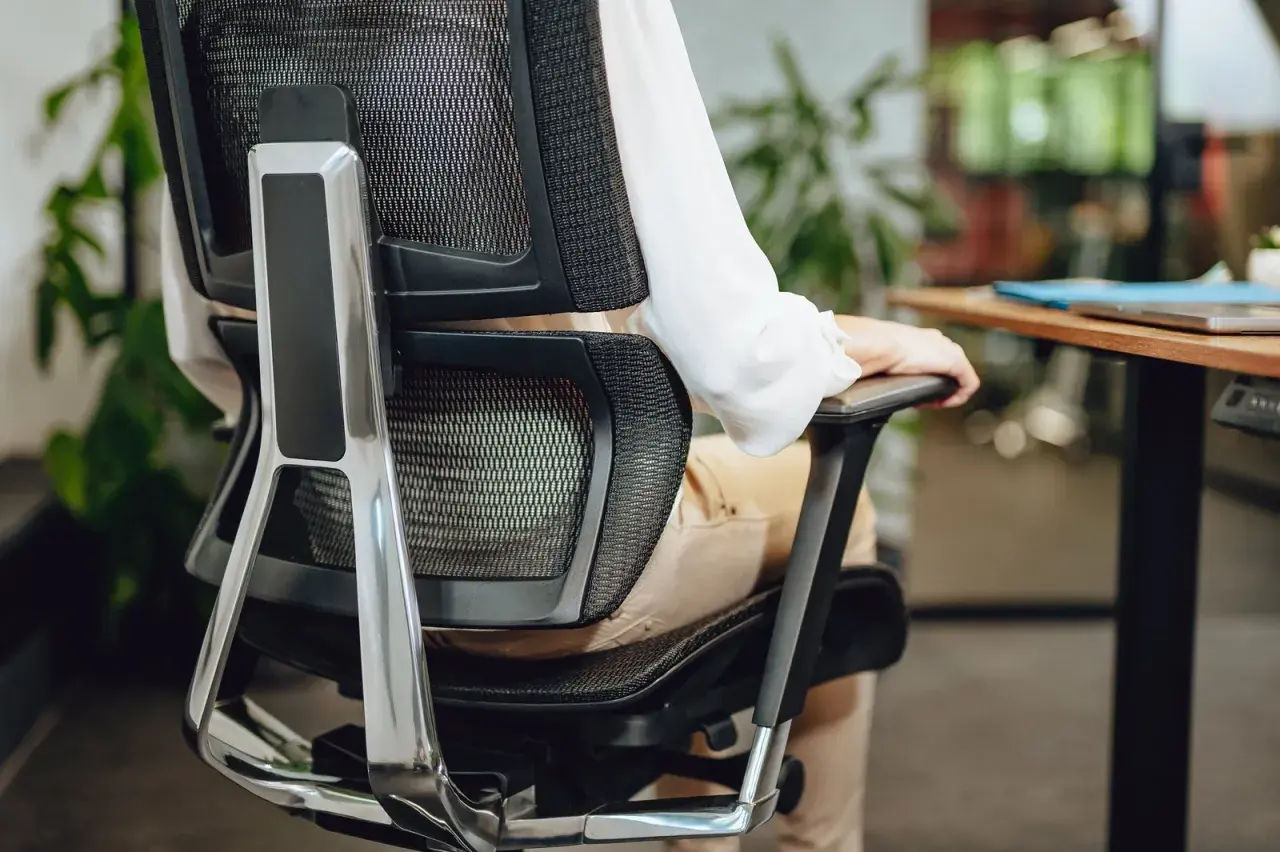 Person in bequemer Sitzposition am Schreibtisch, die Armlehne des Bürostuhls nutzend.