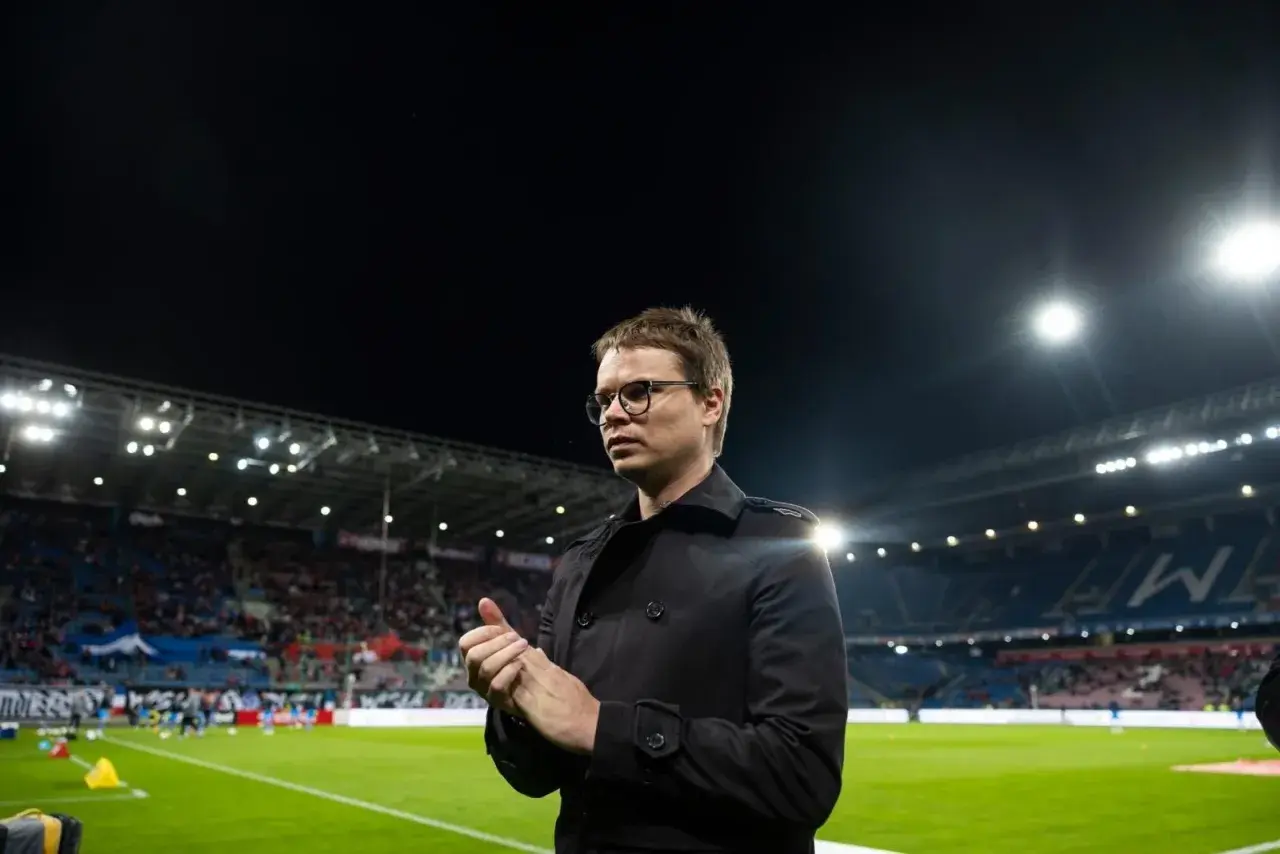 Właściciel Wisły Kraków, w czarnym płaszczu, stoi na murawie stadionu, patrząc w bok.