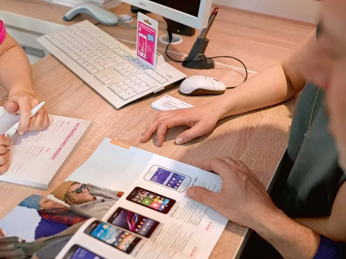 Klienci przeglądają ofertę smartfonów T-Mobile. Na biurku klawiatura, myszka i dokumenty.