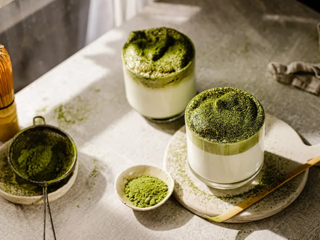 Zwei Gläser cremiger Matcha-Latte, bestreut mit feinem Pulver. Die Zubereitung mit Chasen und Sieb deutet auf die belebende matcha wirkung hin.