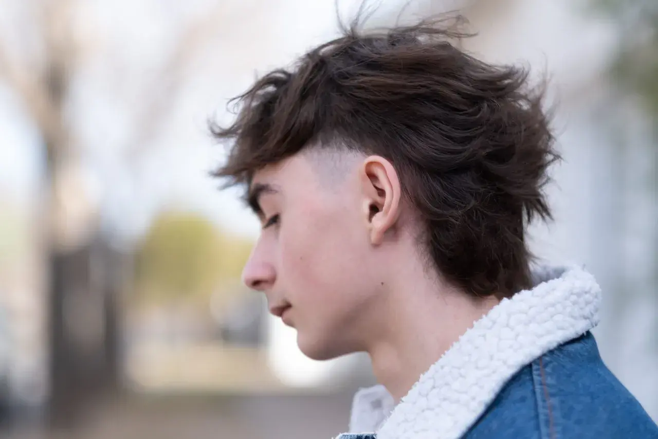 Junger Mann mit modernen Mullets, der nachdenklich zur Seite blickt. Sein Haar ist dunkelbraun und zerzaust, die Seiten sind kurz geschoren. Er trägt eine Jeansjacke mit Teddyfellkragen.