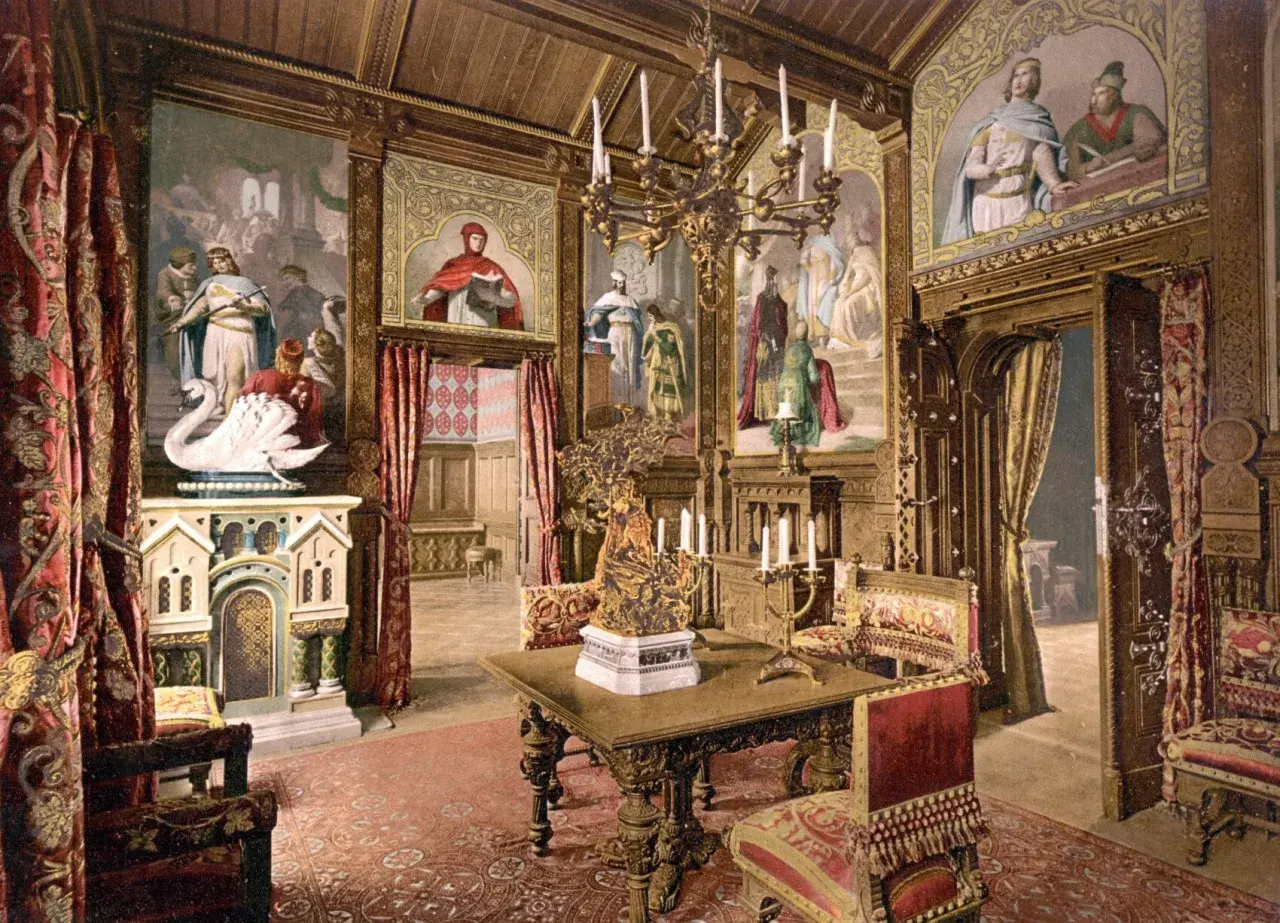 Prunkvoller Salon im Schloss Neuschwanstein innen. Wandmalereien mit Sagenmotiven, ein Kamin mit Schwan, prunkvolle Möbel und ein Kronleuchter schaffen königliches Ambiente.