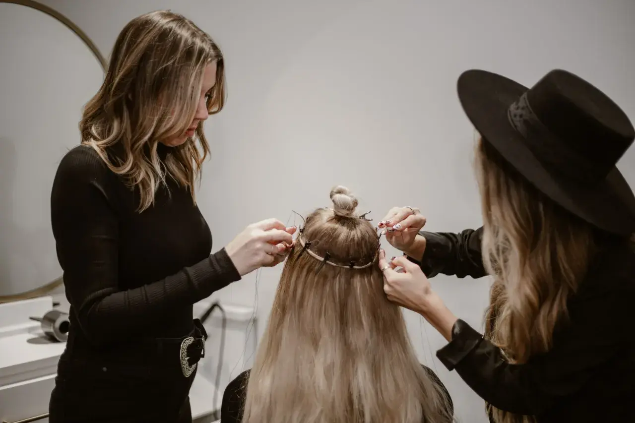 Två personer hjälper till att **slinga håret hemma** med extensions. En med svart polotröja och en med svart hatt arbetar på det långa blonda håret.