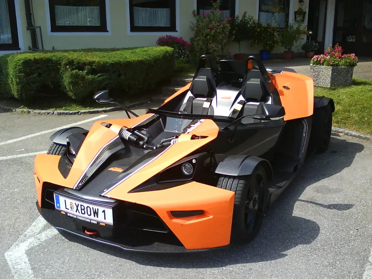 Pomarańczowy KTM X-Bow z czarnymi akcentami i tablicą rejestracyjną "XBOW 1" stoi na parkingu przed budynkiem z zielonym żywopłotem.