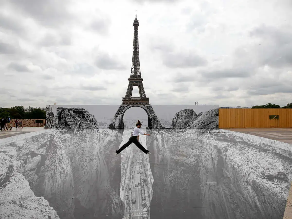 Une femme saute au-dessus d'un gouffre dessiné par un peintre trompe l'oeil connu, avec la Tour Eiffel en arrière-plan.