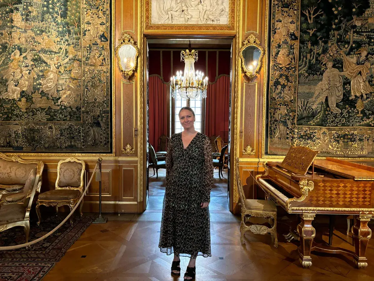 En kvinna står i en sal på Hallwylska museet Stockholm, omgiven av antika gobelänger, en flygel och utsökt inredning.