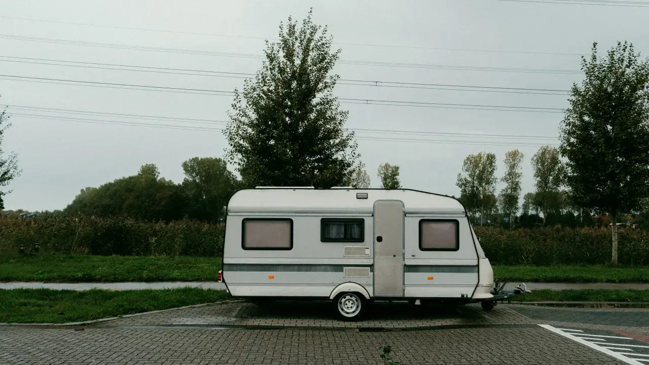 Ein weißer b96 wohnwagen steht auf einem gepflasterten Parkplatz, umgeben von grüner Wiese und Bäumen unter einem grauen Himmel.