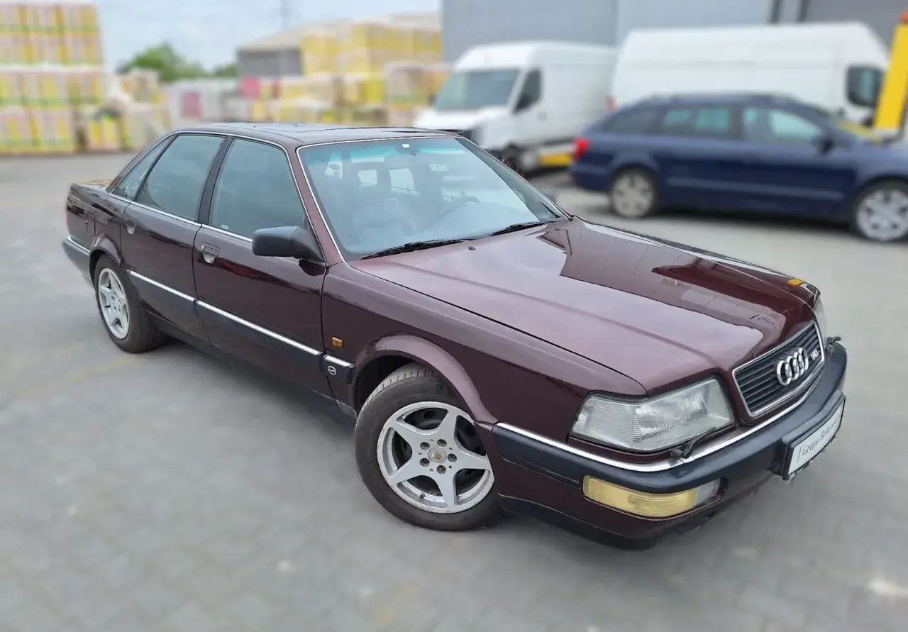 Bordowy Audi V8 z lat 90. na parkingu, w tle białe vany i niebieski kombi.