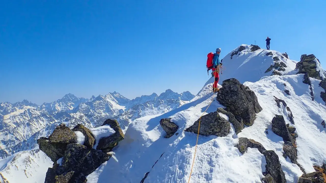 Wspinaczka zimą na Orlą Perć. Dwóch alpinistów na ośnieżonym szczycie, jeden z nich zabezpiecza się liną.