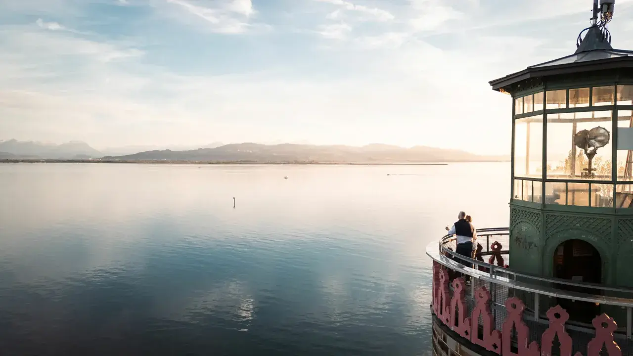 Ein Paar genießt den Sonnenuntergang am Leuchtturm Lindau. Der Bodensee glänzt, Berge im Hintergrund.