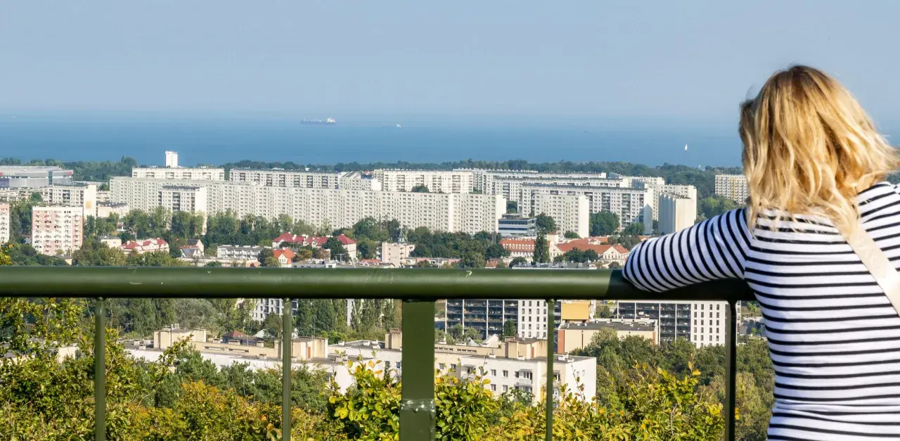 Widok z punktu widokowego w Oliwie na morze i osiedle. Kobieta w bluzce w paski podziwia panoramę.
