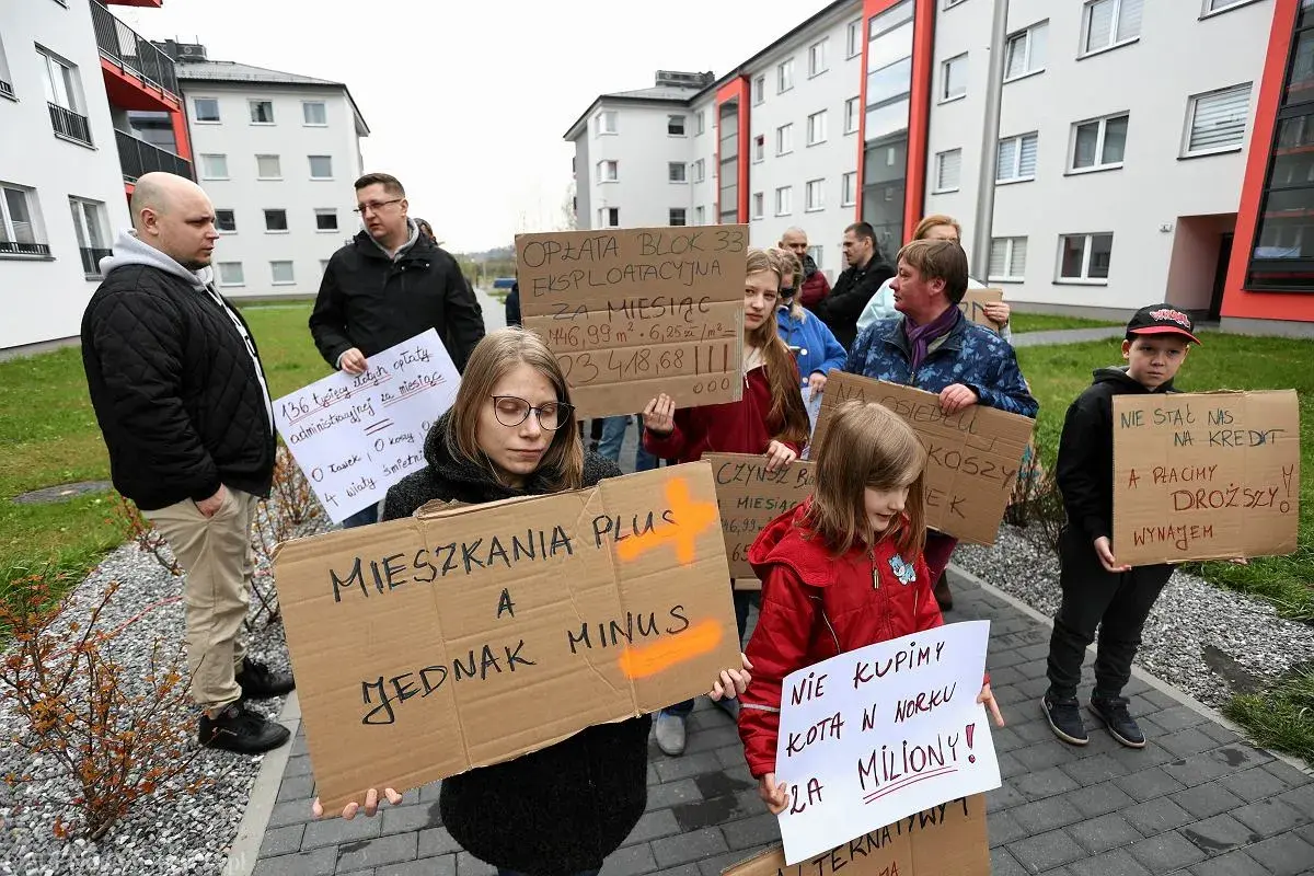 Ludzie protestują pod blokami, trzymając transparenty. Jeden z nich głosi "Mieszkania Plus, a jednak minus".