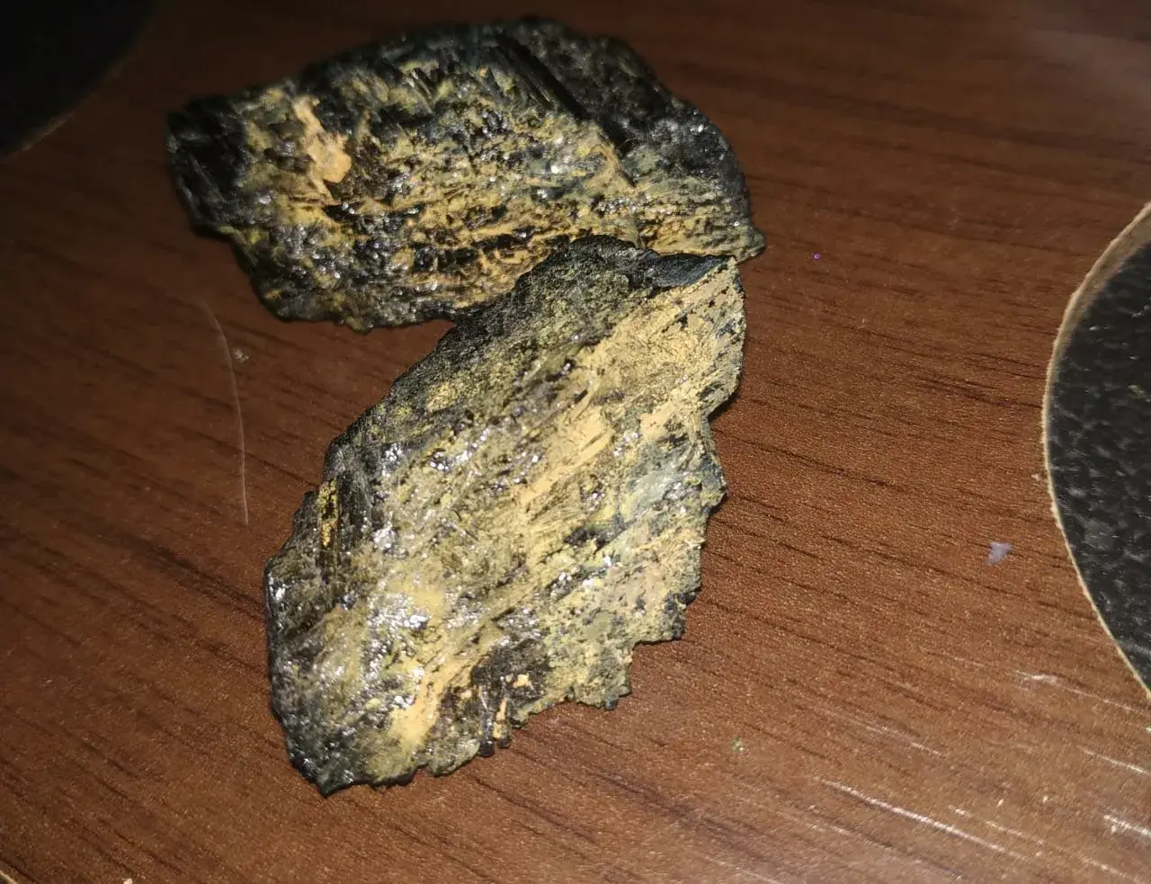 Dwa kawałki czarnego turmalinu, które pękły, pokryte żółtym nalotem, leżą na drewnianym blacie.