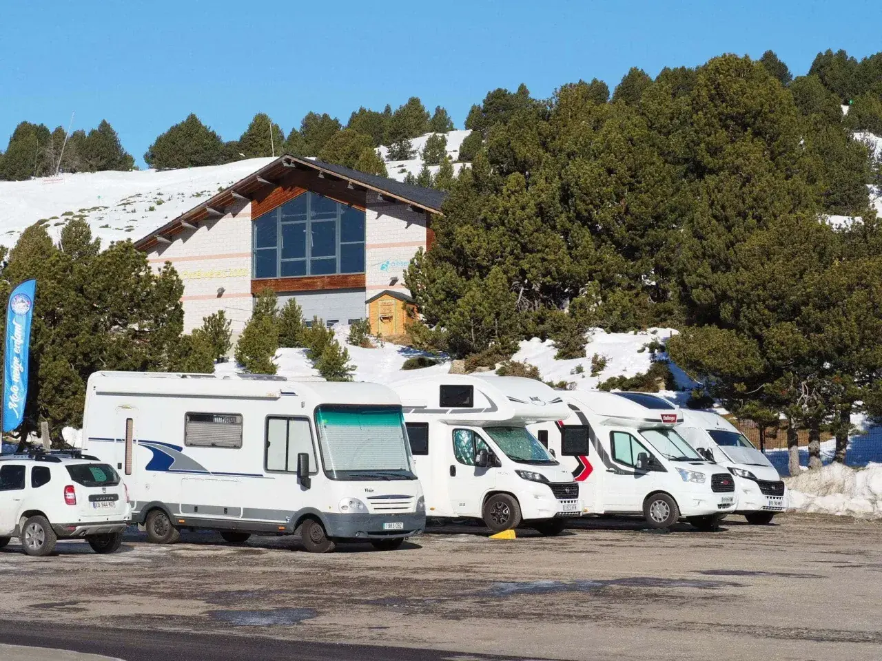 Stationnement de camping-cars dans une aire de camping car Andorre, avec un bâtiment et des pins enneigés en arrière-plan.