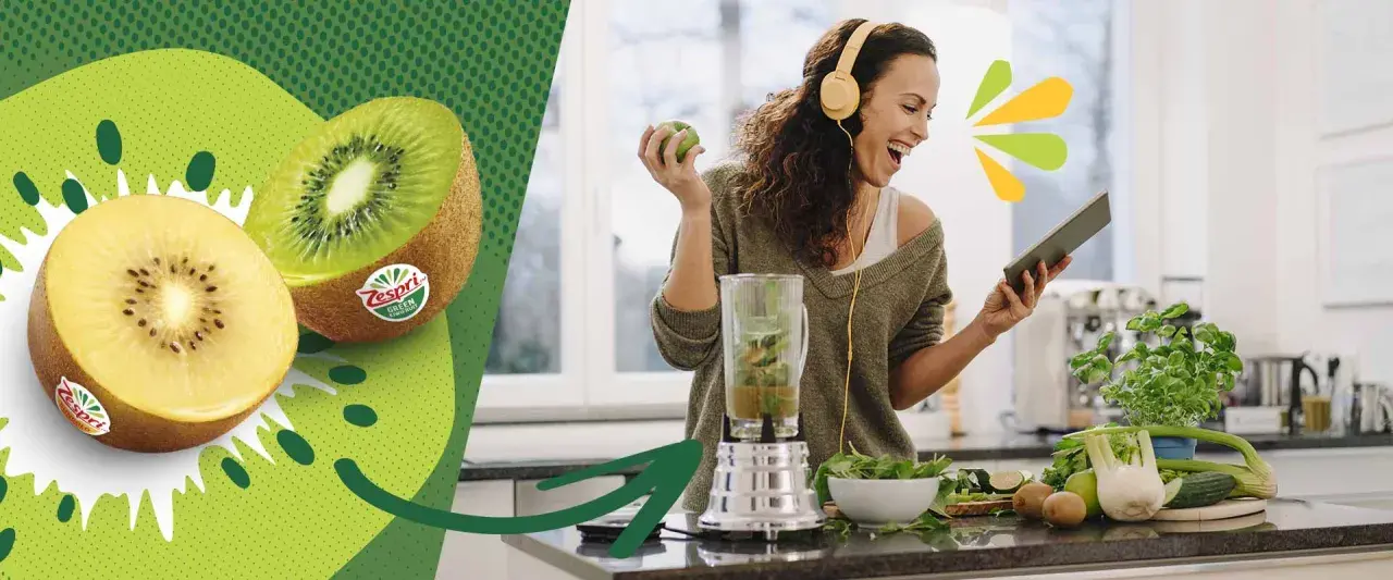 Glad kvinna med hörlurar gör smoothie med kiwi nyttigt. Två halverade kiwifrukter syns till vänster.