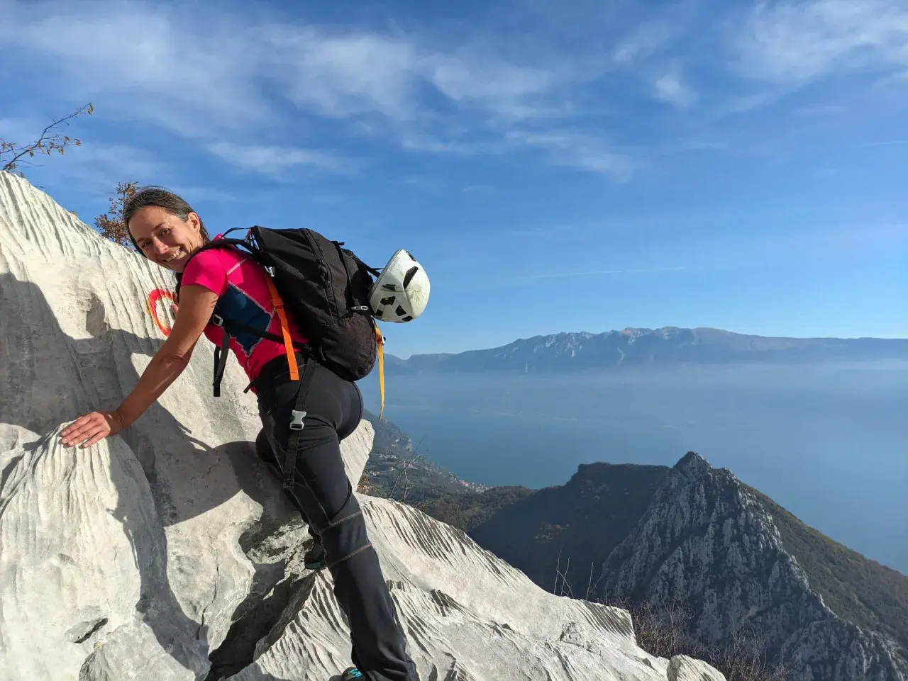Donna sorridente su roccia con zaino e casco, vista lago e montagne. Un facile sentiero sul monte Pizzocolo.