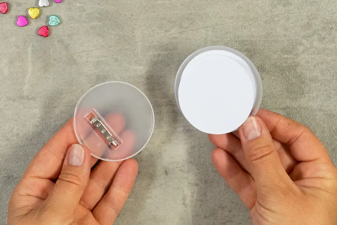 Zwei Hände halten Teile für das **button basteln**: eine mit einer transparenten Rückseite und Nadel, die andere mit einem weißen Kreis.