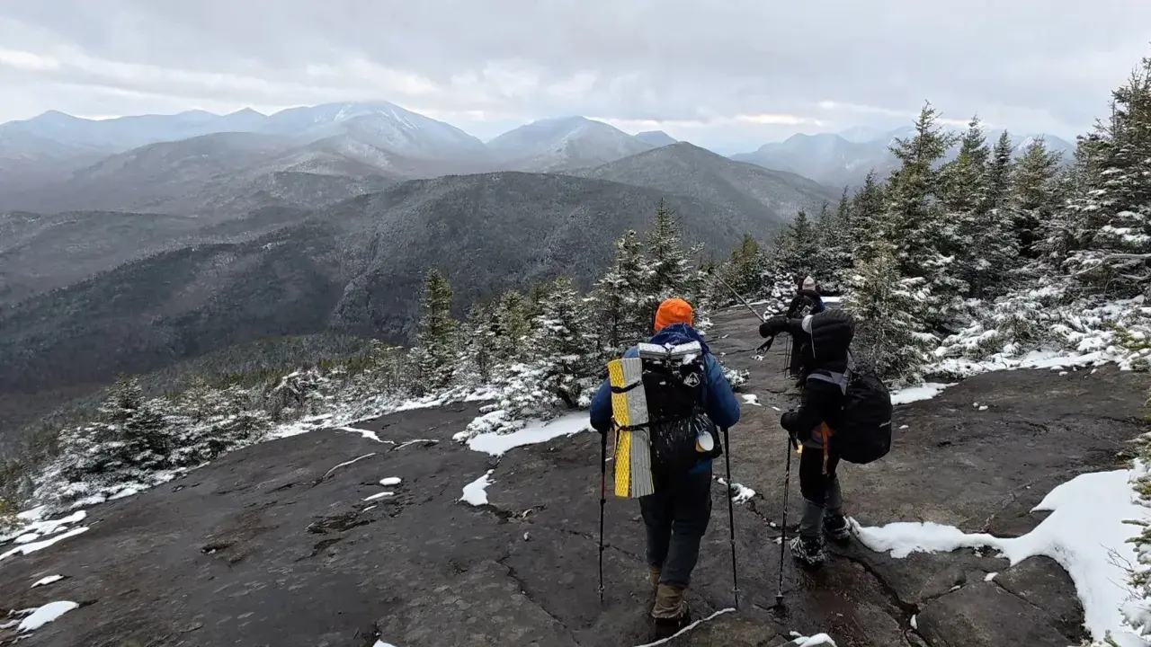 Hikers trek across a snowy mountain ridge, leaving no hiker trash behind. The vast, misty mountain range stretches into the distance under a cloudy sky.