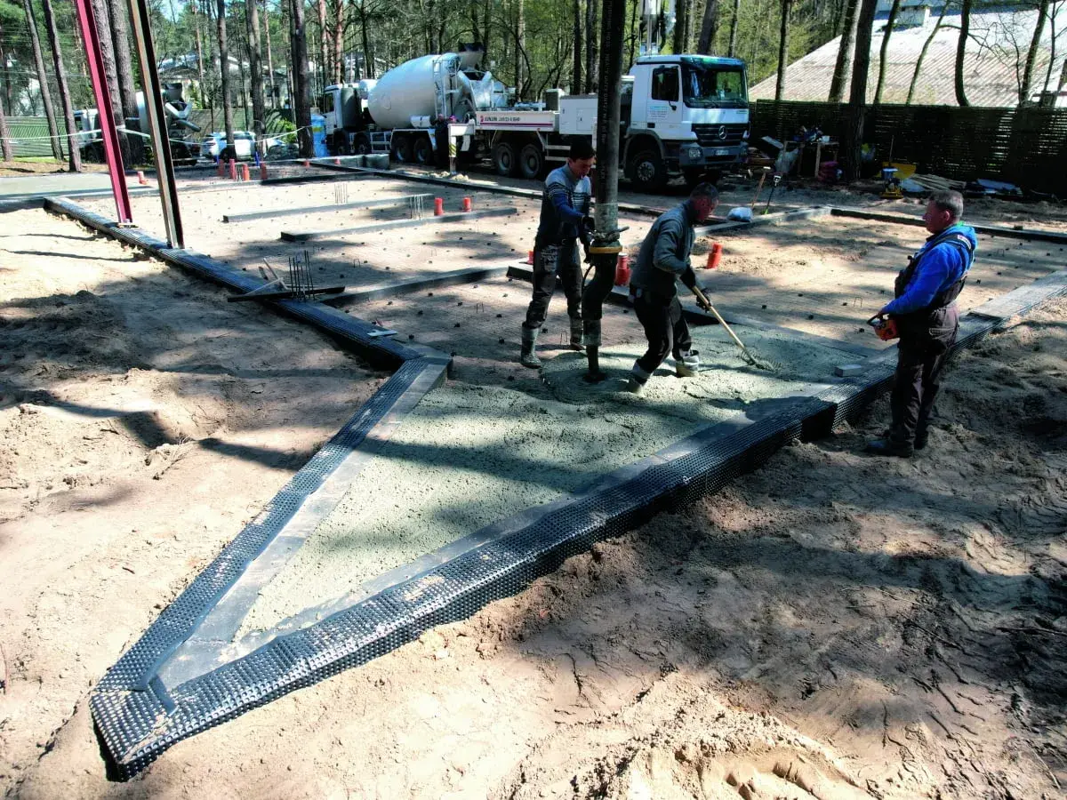 Pracownicy wylewają beton na budowie, przygotowując podłoże pod posadzkę. Widać ciężarówkę z mieszanką betonową i sprzęt budowlany.