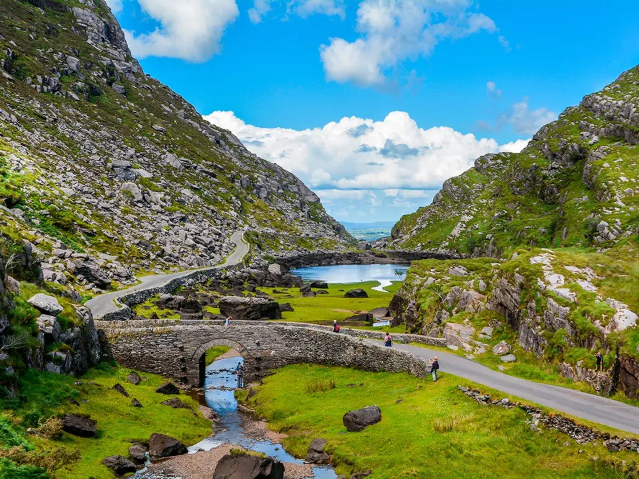 Eine malerische **irland landschaft** mit einem steinernen Bogenbrücken über einem Bach, der in einen See mündet, umgeben von grünen Hügeln und felsigen Bergen.