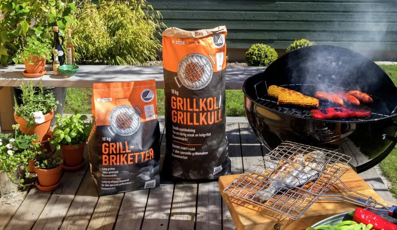 Två säckar med grillkol eller briketter står redo för grillkvällen. Majskolvar, korvar och paprika ryker på grillen.