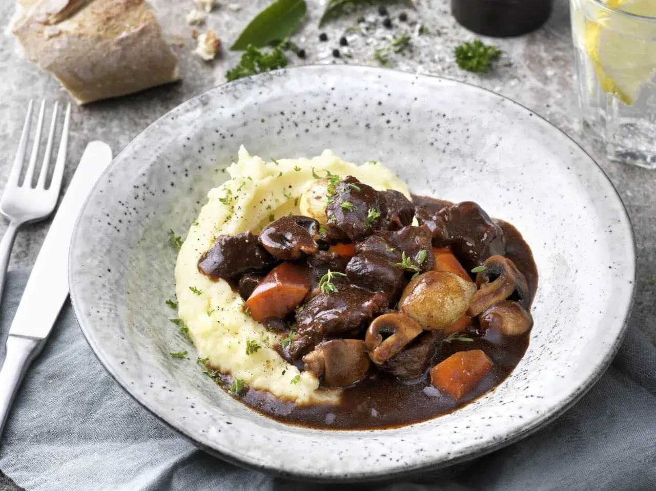 En mustig boeuf bourguignon med potatismos, champinjoner och morötter. Serveras i en rustik skål med en bit bröd och ett glas vatten.