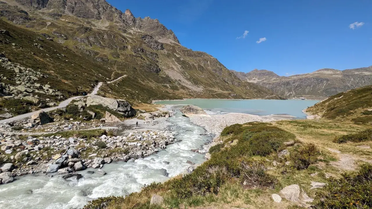 Berglandschaft mit türkisfarbenem Wasser des Silvretta Stausees, der von einem Fluss gespeist wird. Wanderweg schlängelt sich den Hang hinauf.